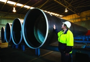 A worker in hi-vis and hard hat beside three large-diameter steel pipes inside a plant