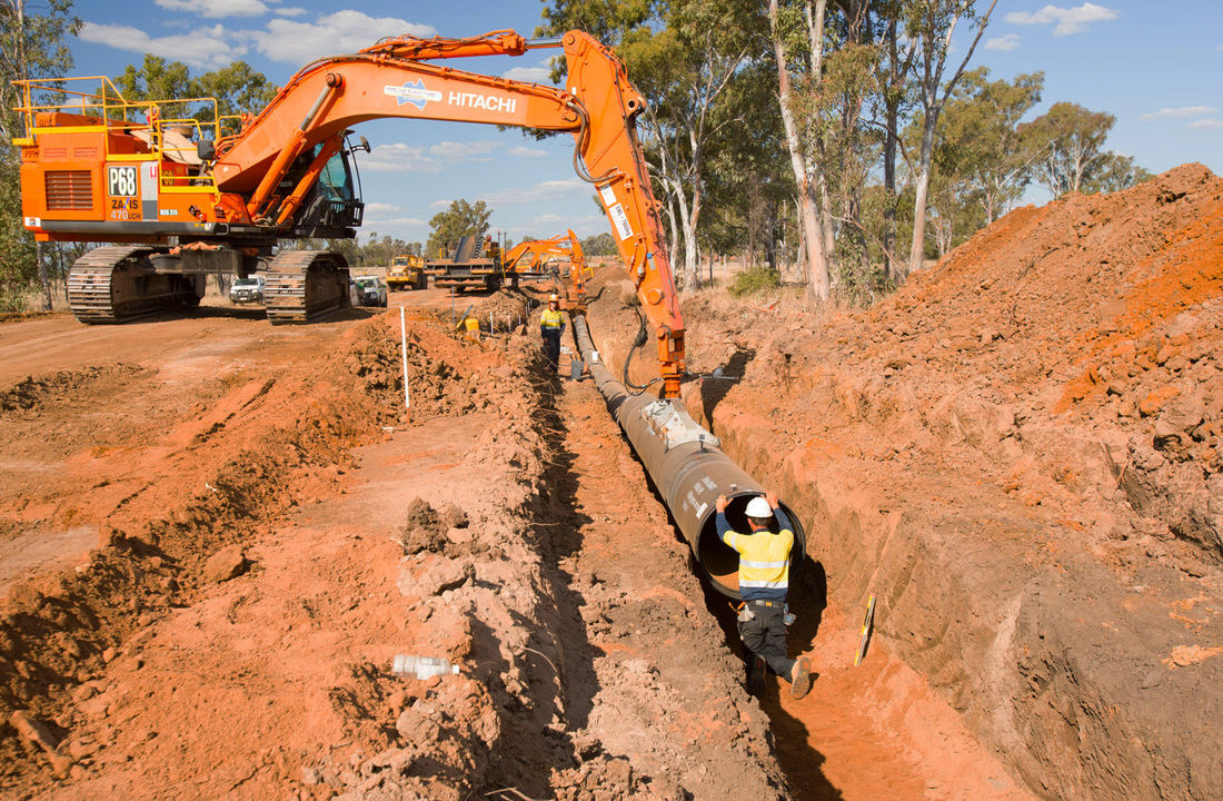 An excavator lowers coated steel pipe into a red-earth trench while a worker in hi-vis guides it into place